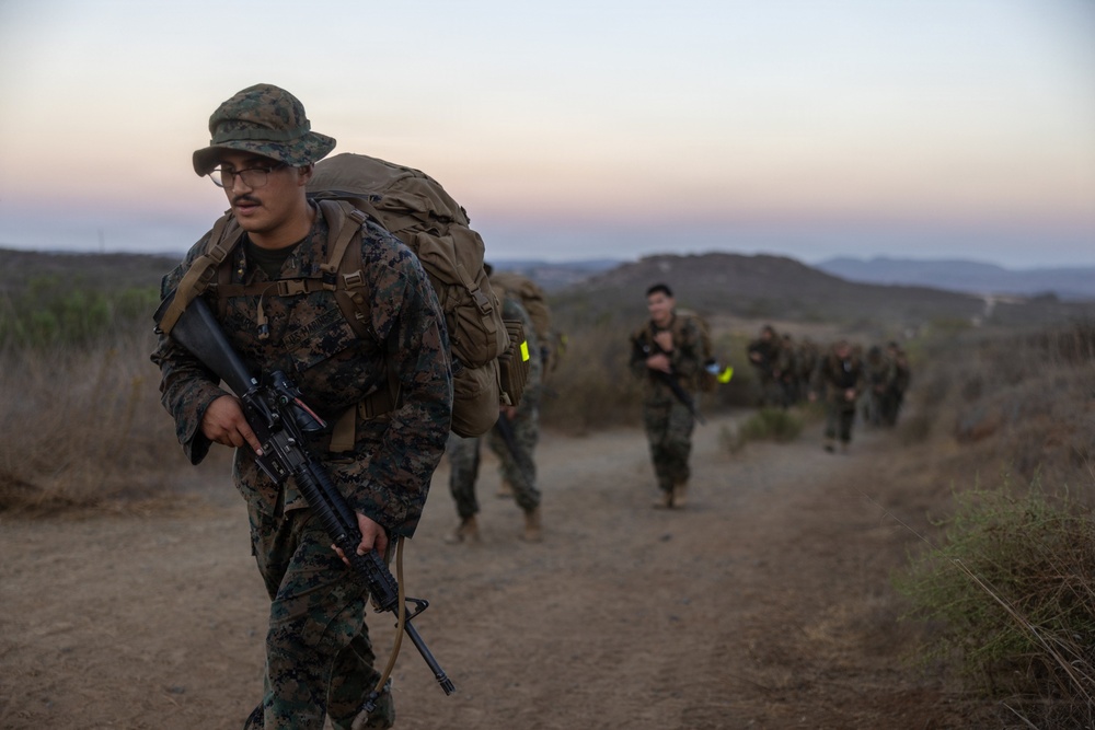 A column of military personnel in MARPAT (Marine pattern) camouflage uniforms conduct a tactical foot march or 'hump' along a dirt trail in a hilly, semi-arid terrain at dusk or dawn. The lead Marine carries an M16/M4-style rifle and wears a boonie cover while bearing a large rucksack. Multiple service members follow in a column formation stretching into the background.