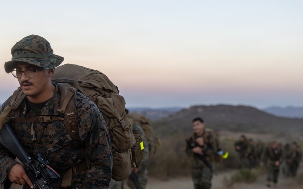 A column of military personnel in MARPAT (Marine pattern) camouflage uniforms conduct a tactical foot march or 'hump' along a dirt trail in a hilly, semi-arid terrain at dusk or dawn. The lead Marine carries an M16/M4-style rifle and wears a boonie cover while bearing a large rucksack. Multiple service members follow in a column formation stretching into the background.