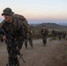 A column of military personnel in MARPAT (Marine pattern) camouflage uniforms conduct a tactical foot march or 'hump' along a dirt trail in a hilly, semi-arid terrain at dusk or dawn. The lead Marine carries an M16/M4-style rifle and wears a boonie cover while bearing a large rucksack. Multiple service members follow in a column formation stretching into the background.