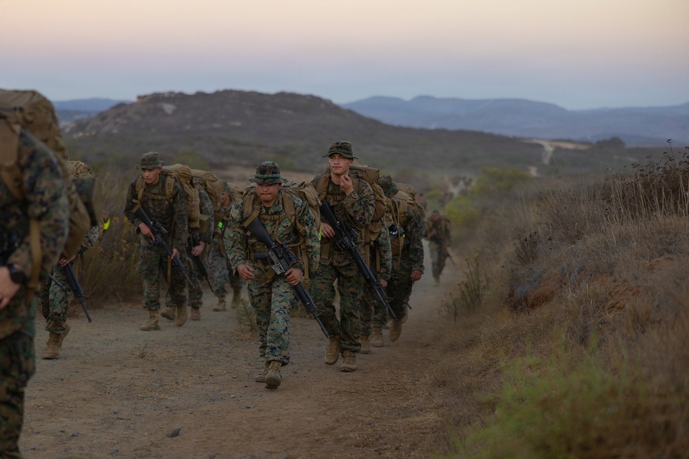 A column of military personnel in camouflage uniforms conducts a loaded ruck march along a dirt trail in a hilly, arid terrain. The soldiers are carrying large backpacks and rifles, moving in formation toward the camera. The setting appears to be a training environment, likely during early morning or dusk based on the soft, low light conditions.