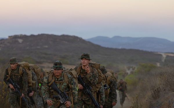 A column of military personnel in camouflage uniforms conducts a loaded ruck march along a dirt trail in a hilly, arid terrain. The soldiers are carrying large backpacks and rifles, moving in formation toward the camera. The setting appears to be a training environment, likely during early morning or dusk based on the soft, low light conditions.