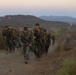 A column of military personnel in camouflage uniforms conducts a loaded ruck march along a dirt trail in a hilly, arid terrain. The soldiers are carrying large backpacks and rifles, moving in formation toward the camera. The setting appears to be a training environment, likely during early morning or dusk based on the soft, low light conditions.