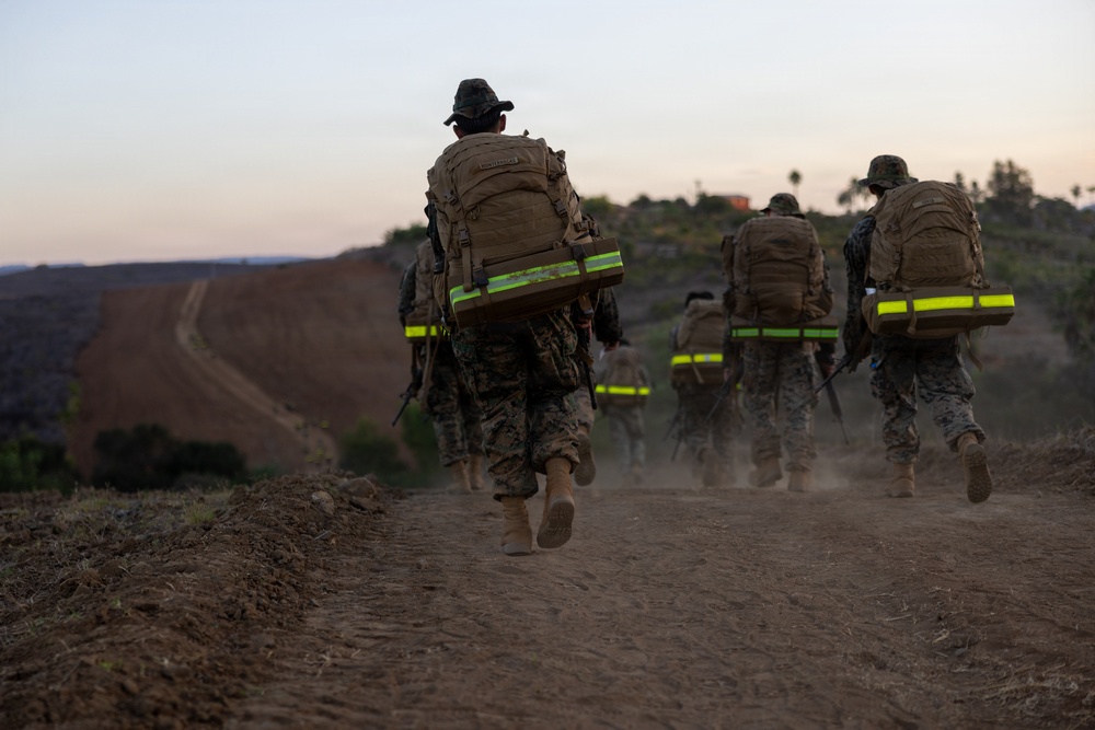 A group of military personnel in camouflage uniforms are conducting a ruck march along a dirt trail at dusk or dawn. They are carrying large tactical backpacks equipped with high-visibility yellow-green reflective strips, likely for safety during low-light conditions. The terrain appears arid and hilly, consistent with a training environment.