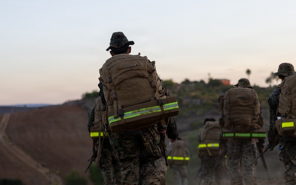 A group of military personnel in camouflage uniforms are conducting a ruck march along a dirt trail at dusk or dawn. They are carrying large tactical backpacks equipped with high-visibility yellow-green reflective strips, likely for safety during low-light conditions. The terrain appears arid and hilly, consistent with a training environment.