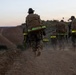 A group of military personnel in camouflage uniforms are conducting a ruck march along a dirt trail at dusk or dawn. They are carrying large tactical backpacks equipped with high-visibility yellow-green reflective strips, likely for safety during low-light conditions. The terrain appears arid and hilly, consistent with a training environment.