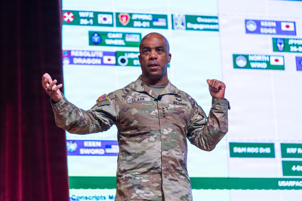 A U.S. Army officer with the name tag 'CLARK' and three stars on his chest (indicating the rank of Lieutenant General) is delivering a presentation on stage. He is gesturing with both hands while speaking in front of a large projected slide showing a military exercise schedule. The slide references various joint military exercises including 'Keen Sword,' 'Resolute Dragon,' 'North Wind,' and 'Keen Edge,' with flags indicating multinational participation.
