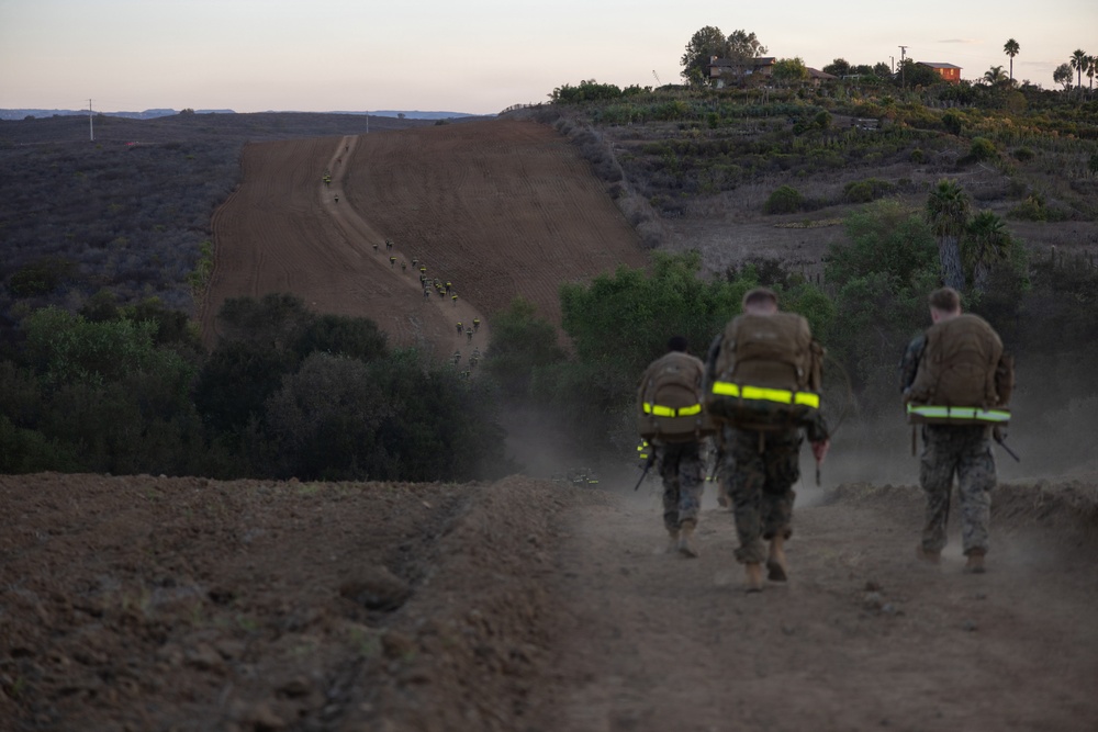 Three military personnel in combat gear with reflective safety strips on their backpacks are hiking along a dusty dirt trail at dusk or dawn. In the distance, a larger group of similarly equipped personnel can be seen further along the same trail. The setting appears to be a hilly, semi-arid training area with vegetation and a structure visible on the hilltop.