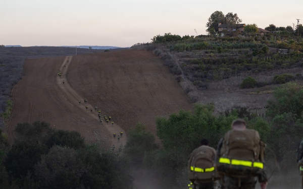 Three military personnel in combat gear with reflective safety strips on their backpacks are hiking along a dusty dirt trail at dusk or dawn. In the distance, a larger group of similarly equipped personnel can be seen further along the same trail. The setting appears to be a hilly, semi-arid training area with vegetation and a structure visible on the hilltop.