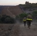 Three military personnel in combat gear with reflective safety strips on their backpacks are hiking along a dusty dirt trail at dusk or dawn. In the distance, a larger group of similarly equipped personnel can be seen further along the same trail. The setting appears to be a hilly, semi-arid training area with vegetation and a structure visible on the hilltop.