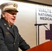 A senior fire department official in dress uniform is speaking at a podium during a formal event at Walter Reed National Military Medical Center. The individual is wearing a white-topped chief's cap and a dark navy dress uniform with a Raleigh, N.C. fire department patch. The setting appears to be an indoor ceremony or official presentation.