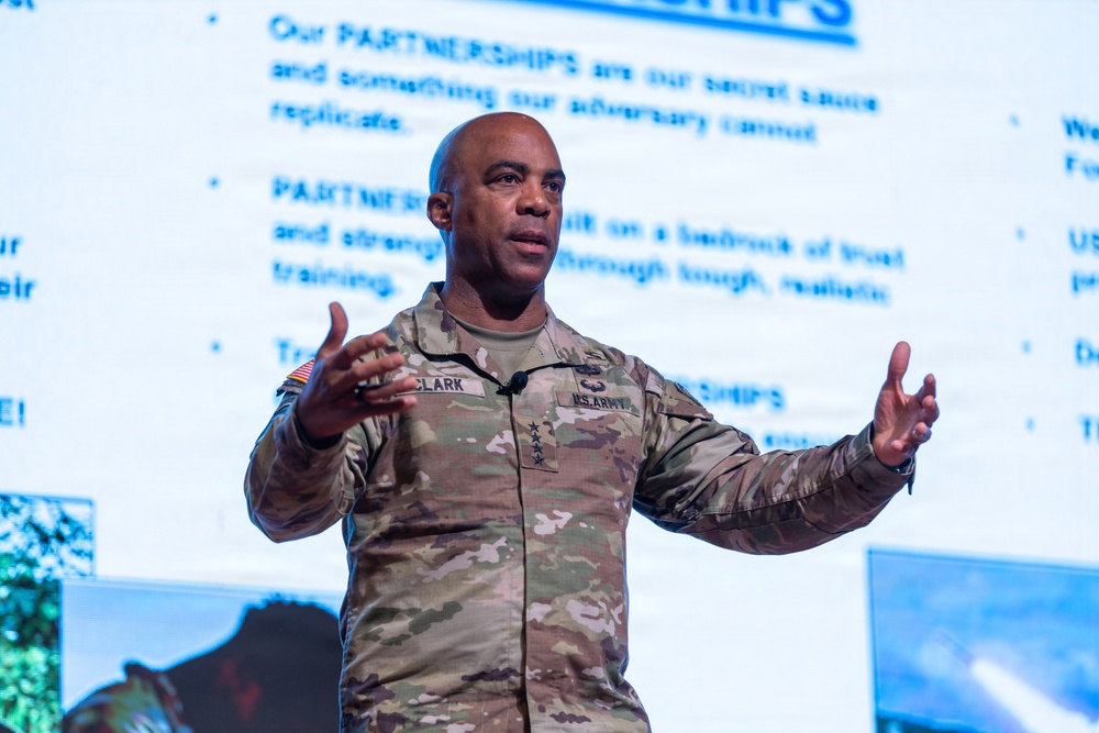 A U.S. Army officer with the name tag 'CLARK' and four-star general rank insignia is delivering a presentation in front of a projected slide about 'Partnerships.' He is gesturing with both hands raised, appearing engaged in active speech. The slide behind him emphasizes that partnerships are a strategic advantage that adversaries cannot replicate.