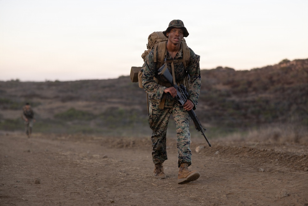 A U.S. Marine in full combat gear is conducting a ruck march across arid, open terrain at dusk or dawn. The Marine is carrying a large pack and holding a rifle while moving at a brisk pace along a dirt trail. A second service member is visible in the background, similarly equipped.