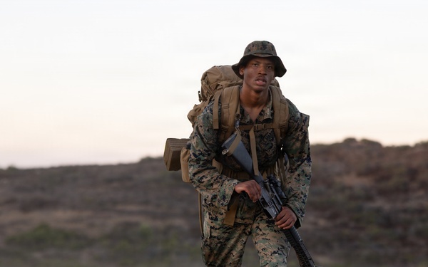 A U.S. Marine in full combat gear is conducting a ruck march across arid, open terrain at dusk or dawn. The Marine is carrying a large pack and holding a rifle while moving at a brisk pace along a dirt trail. A second service member is visible in the background, similarly equipped.