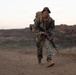 A U.S. Marine in full combat gear is conducting a ruck march across arid, open terrain at dusk or dawn. The Marine is carrying a large pack and holding a rifle while moving at a brisk pace along a dirt trail. A second service member is visible in the background, similarly equipped.