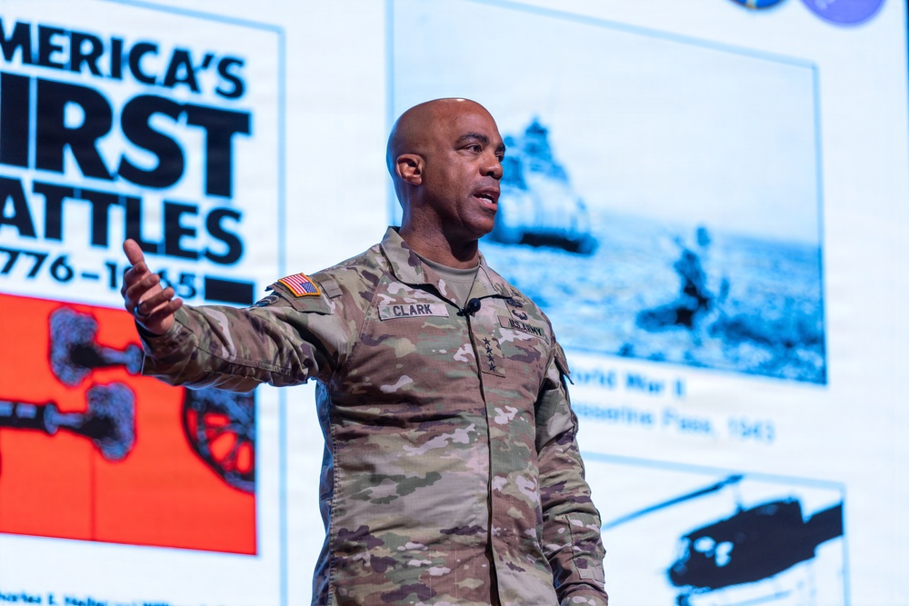 A U.S. Army general officer with the name tag 'CLARK' and four-star rank insignia is delivering a presentation on stage. He is wearing the Army Combat Uniform (ACU) with a U.S. flag patch and 'US ARMY' tape visible. The background projection displays a slide titled 'America's First Battles 1776-1965' with historical military imagery.