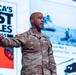 A U.S. Army general officer with the name tag 'CLARK' and four-star rank insignia is delivering a presentation on stage. He is wearing the Army Combat Uniform (ACU) with a U.S. flag patch and 'US ARMY' tape visible. The background projection displays a slide titled 'America's First Battles 1776-1965' with historical military imagery.