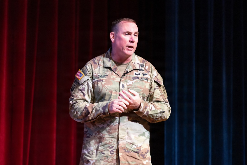 A U.S. Army soldier named Schmidt, wearing an OCP (Operational Camouflage Pattern) uniform, is speaking or presenting on a stage. He appears to be addressing an audience, with his hands clasped together in front of him. The backdrop consists of red and blue curtains, suggesting a formal auditorium or theater setting.