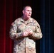 A U.S. Army soldier named Schmidt, wearing an OCP (Operational Camouflage Pattern) uniform, is speaking or presenting on a stage. He appears to be addressing an audience, with his hands clasped together in front of him. The backdrop consists of red and blue curtains, suggesting a formal auditorium or theater setting.