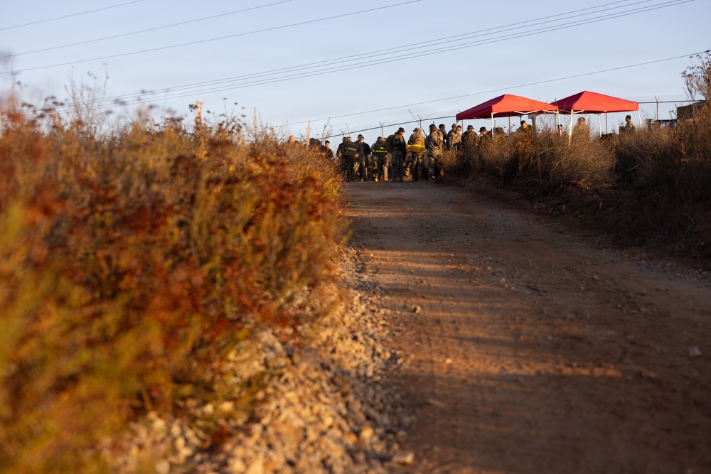 A dirt/gravel road leads toward a group of uniformed personnel gathered near two red canopy tents at what appears to be an outdoor staging or command area. The scene is photographed during golden hour lighting, with dry autumn vegetation flanking the road on both sides. Power lines run overhead parallel to the road.