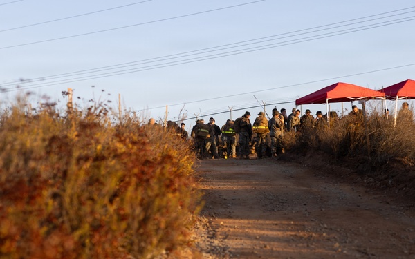 A dirt/gravel road leads toward a group of uniformed personnel gathered near two red canopy tents at what appears to be an outdoor staging or command area. The scene is photographed during golden hour lighting, with dry autumn vegetation flanking the road on both sides. Power lines run overhead parallel to the road.