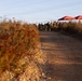 A dirt/gravel road leads toward a group of uniformed personnel gathered near two red canopy tents at what appears to be an outdoor staging or command area. The scene is photographed during golden hour lighting, with dry autumn vegetation flanking the road on both sides. Power lines run overhead parallel to the road.