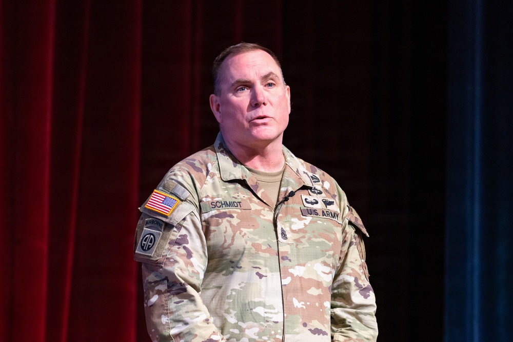 A U.S. Army soldier named Schmidt, affiliated with the 82nd Airborne Division, is standing and speaking at what appears to be a formal military presentation or event. He is wearing an OCP (Operational Camouflage Pattern) uniform with visible rank insignia of a Senior Non-Commissioned Officer. The setting appears to be a stage with red and blue curtains in the background.