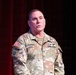 A U.S. Army soldier named Schmidt, affiliated with the 82nd Airborne Division, is standing and speaking at what appears to be a formal military presentation or event. He is wearing an OCP (Operational Camouflage Pattern) uniform with visible rank insignia of a Senior Non-Commissioned Officer. The setting appears to be a stage with red and blue curtains in the background.