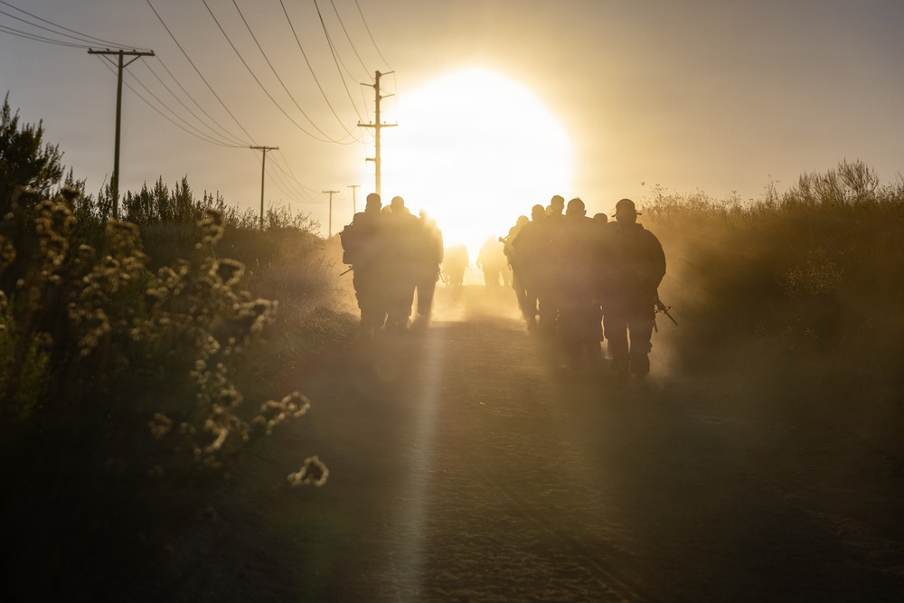 A group of armed personnel, appearing to be military or law enforcement, march in silhouette along a rural dirt road toward a large, glowing sun on the horizon. The scene is backlit with intense golden light, creating dramatic lens flare and dust haze. Utility poles with power lines line the road, and low scrub vegetation borders both sides.