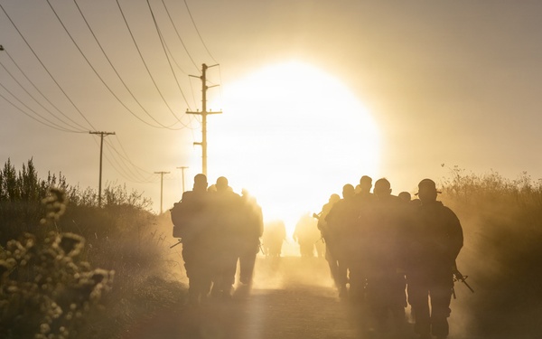 A group of armed personnel, appearing to be military or law enforcement, march in silhouette along a rural dirt road toward a large, glowing sun on the horizon. The scene is backlit with intense golden light, creating dramatic lens flare and dust haze. Utility poles with power lines line the road, and low scrub vegetation borders both sides.