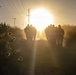 A group of armed personnel, appearing to be military or law enforcement, march in silhouette along a rural dirt road toward a large, glowing sun on the horizon. The scene is backlit with intense golden light, creating dramatic lens flare and dust haze. Utility poles with power lines line the road, and low scrub vegetation borders both sides.