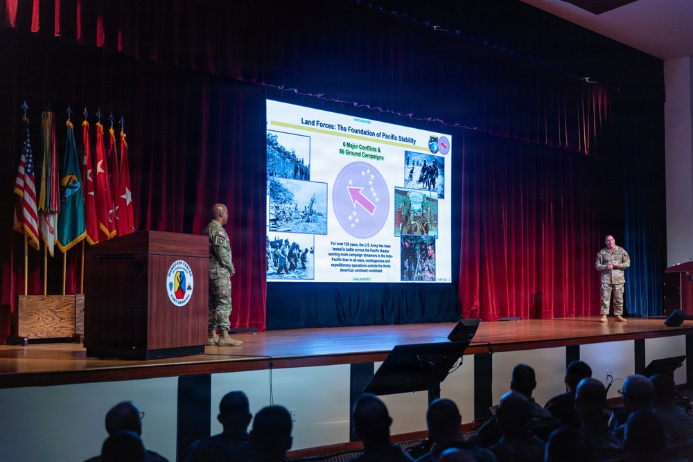 A military briefing or presentation is taking place in an auditorium, with two uniformed soldiers on stage presenting a slide titled 'Land Forces: The Foundation of Pacific stability.' The presentation references '6 Major Conflicts &amp; 86 Ground Campaigns' and features what appears to be a U.S. Army Pacific (USARPAC) unit insignia. An audience of military and civilian personnel is seated in the foreground observing the presentation.
