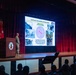 A military briefing or presentation is taking place in an auditorium, with two uniformed soldiers on stage presenting a slide titled 'Land Forces: The Foundation of Pacific stability.' The presentation references '6 Major Conflicts &amp; 86 Ground Campaigns' and features what appears to be a U.S. Army Pacific (USARPAC) unit insignia. An audience of military and civilian personnel is seated in the foreground observing the presentation.