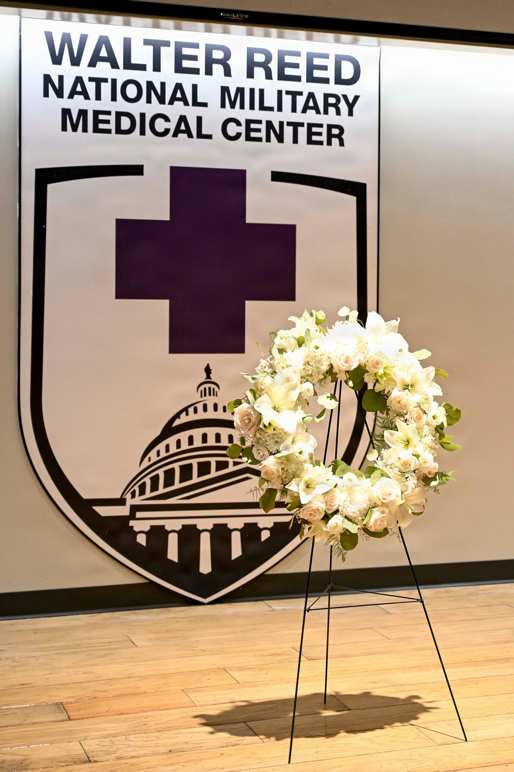 The image shows a formal memorial or ceremonial setting at Walter Reed National Military Medical Center. A large institutional banner displaying the center's shield logo — featuring a purple cross and the U.S. Capitol dome — serves as a backdrop. In the foreground, an elegant white floral wreath composed of roses, lilies, and greenery is displayed on a black metal easel stand.