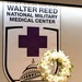 The image shows a formal memorial or ceremonial setting at Walter Reed National Military Medical Center. A large institutional banner displaying the center's shield logo — featuring a purple cross and the U.S. Capitol dome — serves as a backdrop. In the foreground, an elegant white floral wreath composed of roses, lilies, and greenery is displayed on a black metal easel stand.