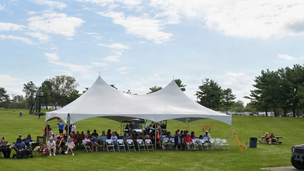 An outdoor gathering is taking place under a large white double-peak event tent on a green lawn. A crowd of approximately 30-50 people are seated in white folding chairs beneath the tent, suggesting a formal or semi-formal community event. The setting appears to be a park or recreational facility with trees, open fields, and what appears to be a watchtower or observation structure in the background.