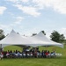 An outdoor gathering is taking place under a large white double-peak event tent on a green lawn. A crowd of approximately 30-50 people are seated in white folding chairs beneath the tent, suggesting a formal or semi-formal community event. The setting appears to be a park or recreational facility with trees, open fields, and what appears to be a watchtower or observation structure in the background.