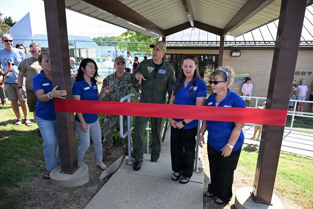 A ribbon-cutting ceremony is taking place at what appears to be a military base Community Recreation facility. Several individuals, including military personnel in uniform and civilians wearing blue polo shirts, are gathered around a red ribbon stretched across an entrance. A crowd of onlookers is visible in the background near what appears to be a swimming pool area.