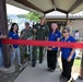 A ribbon-cutting ceremony is taking place at what appears to be a military base Community Recreation facility. Several individuals, including military personnel in uniform and civilians wearing blue polo shirts, are gathered around a red ribbon stretched across an entrance. A crowd of onlookers is visible in the background near what appears to be a swimming pool area.