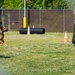 Two military personnel are conducting a military working dog (MWD) training exercise in an enclosed outdoor facility. One soldier is handling a German Shepherd on a leash while the dog lunges aggressively toward a second soldier wearing a protective bite suit vest. The setting includes training obstacles such as a tunnel, traffic cones, and a chain-link fence perimeter.