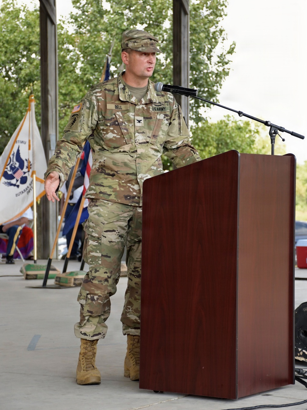 A U.S. Army Colonel named Bell is delivering a speech at a podium equipped with a microphone. He is wearing the Army Combat Uniform (ACU) in MultiCam/OCP pattern with a Colonel's eagle insignia visible on his chest. The setting appears to be an outdoor military ceremony with color guard flags visible in the background.