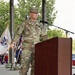 A U.S. Army Colonel named Bell is delivering a speech at a podium equipped with a microphone. He is wearing the Army Combat Uniform (ACU) in MultiCam/OCP pattern with a Colonel's eagle insignia visible on his chest. The setting appears to be an outdoor military ceremony with color guard flags visible in the background.