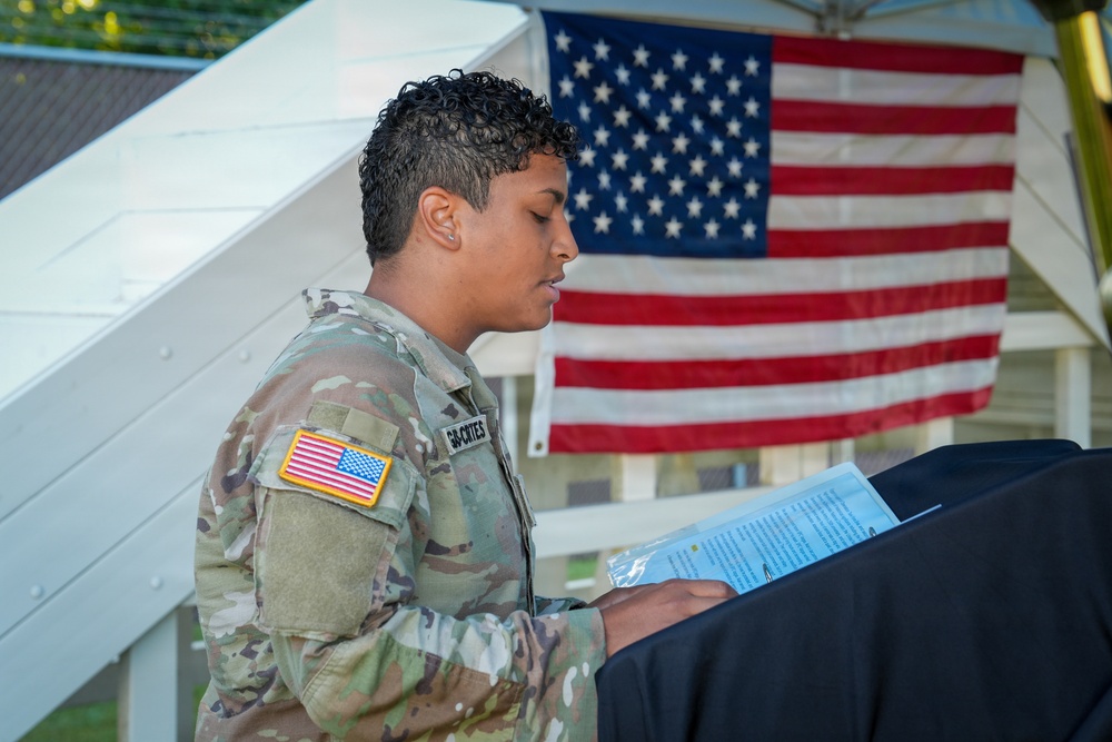 A U.S. Army soldier in Operational Camouflage Pattern (OCP) uniform is reading from a document or program at what appears to be an outdoor military ceremony or event. The soldier has a name tape visible on the uniform and wears a U.S. flag patch on the left sleeve. An American flag is prominently displayed in the background.