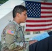 A U.S. Army soldier in Operational Camouflage Pattern (OCP) uniform is reading from a document or program at what appears to be an outdoor military ceremony or event. The soldier has a name tape visible on the uniform and wears a U.S. flag patch on the left sleeve. An American flag is prominently displayed in the background.