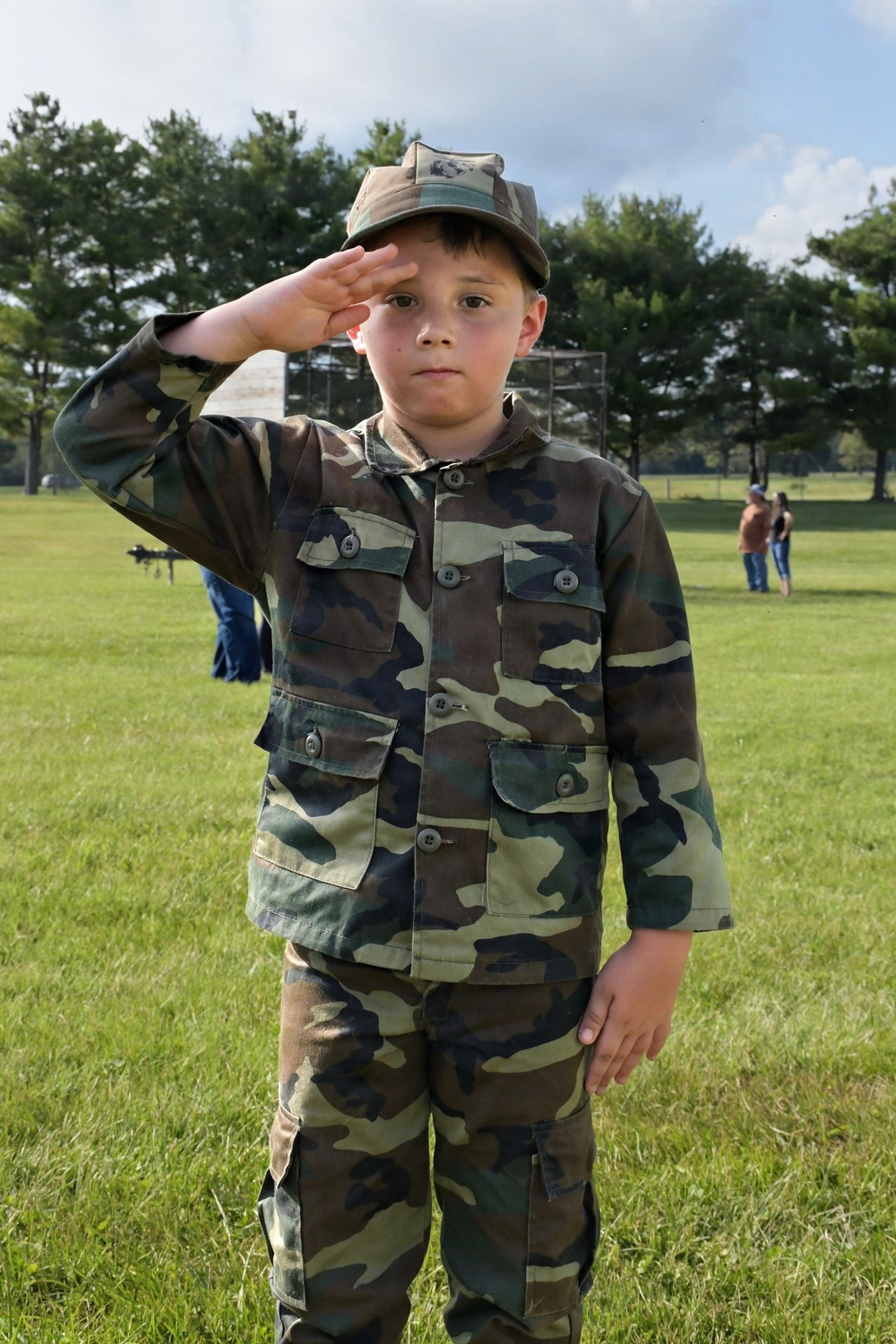 A young child dressed in a full woodland camouflage uniform, including jacket, pants, and matching cap, stands on a grassy field performing a military-style salute. The child has a serious, composed expression directed at the camera. The setting appears to be an outdoor public event or demonstration with other people visible in the background.