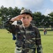 A young child dressed in a full woodland camouflage uniform, including jacket, pants, and matching cap, stands on a grassy field performing a military-style salute. The child has a serious, composed expression directed at the camera. The setting appears to be an outdoor public event or demonstration with other people visible in the background.