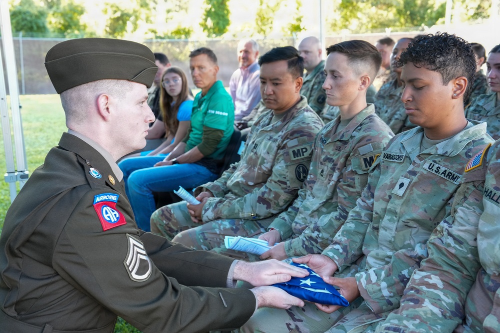 A U.S. Army staff sergeant in dress greens uniform with 82nd Airborne Division patches is presenting a folded American flag to a seated soldier during what appears to be a military ceremony. The ceremony includes both uniformed military personnel in OCP (Operational Camouflage Pattern) uniforms and civilians seated in attendance. The solemn nature of the flag presentation suggests a memorial, retirement, or formal recognition ceremony.