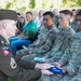 A U.S. Army staff sergeant in dress greens uniform with 82nd Airborne Division patches is presenting a folded American flag to a seated soldier during what appears to be a military ceremony. The ceremony includes both uniformed military personnel in OCP (Operational Camouflage Pattern) uniforms and civilians seated in attendance. The solemn nature of the flag presentation suggests a memorial, retirement, or formal recognition ceremony.