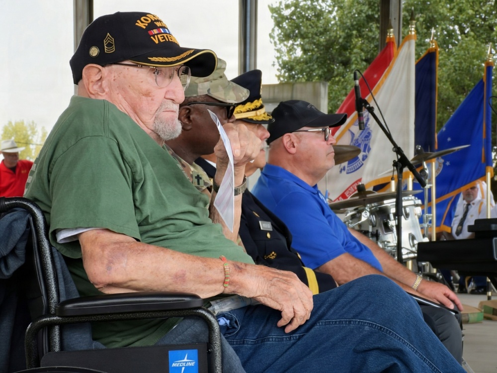 A group of veterans and military personnel are seated at what appears to be an outdoor military ceremony or commemoration event. The most prominent figure is an elderly man in a wheelchair wearing a black 'Korean War Veteran' cap with sergeant major insignia. Behind him are several other attendees including uniformed military officers and civilians, with military flags and a drum kit visible in the background.