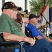 A group of veterans and military personnel are seated at what appears to be an outdoor military ceremony or commemoration event. The most prominent figure is an elderly man in a wheelchair wearing a black 'Korean War Veteran' cap with sergeant major insignia. Behind him are several other attendees including uniformed military officers and civilians, with military flags and a drum kit visible in the background.