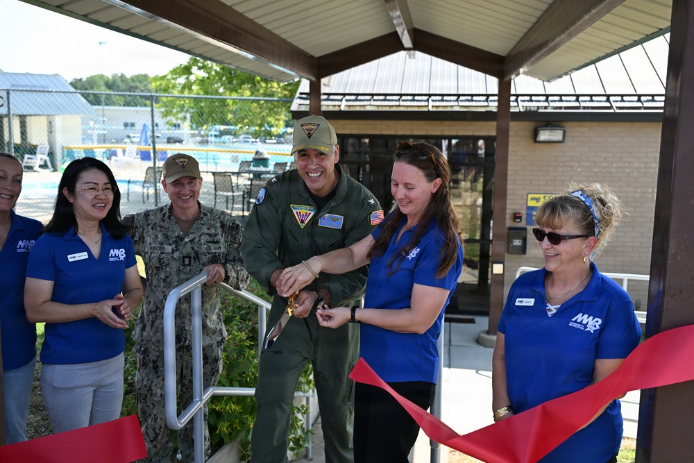 A ribbon-cutting ceremony is taking place at what appears to be a military installation, likely a pool or recreational facility. A military officer in a green flight suit and a woman in a blue MWR (Morale, Welfare, and Recreation) polo shirt are jointly cutting a red ribbon with golden scissors. Several other attendees, including a Navy sailor in camouflage uniform and additional MWR staff members, are present and smiling.